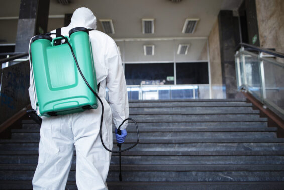 Unrecognizable person in white chemical protection suit with reservoir disinfecting public corridor to stop spreading highly contagious corona virus.